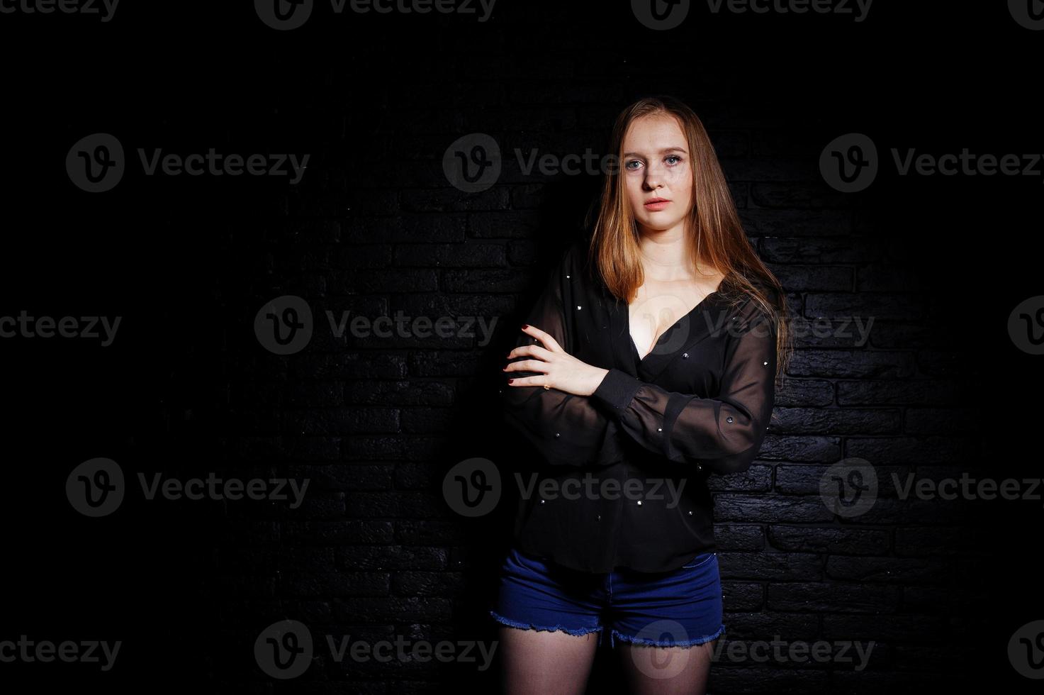 Studio shot of brunette girl in black blouse with bra and shorts against black brick wall. photo