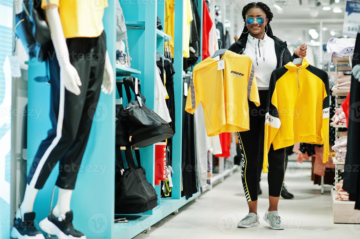 mujeres afroamericanas en chándales y gafas de sol comprando en el centro comercial de ropa deportiva contra estantes. ella elige camiseta amarilla. tema de la tienda de deportes. foto