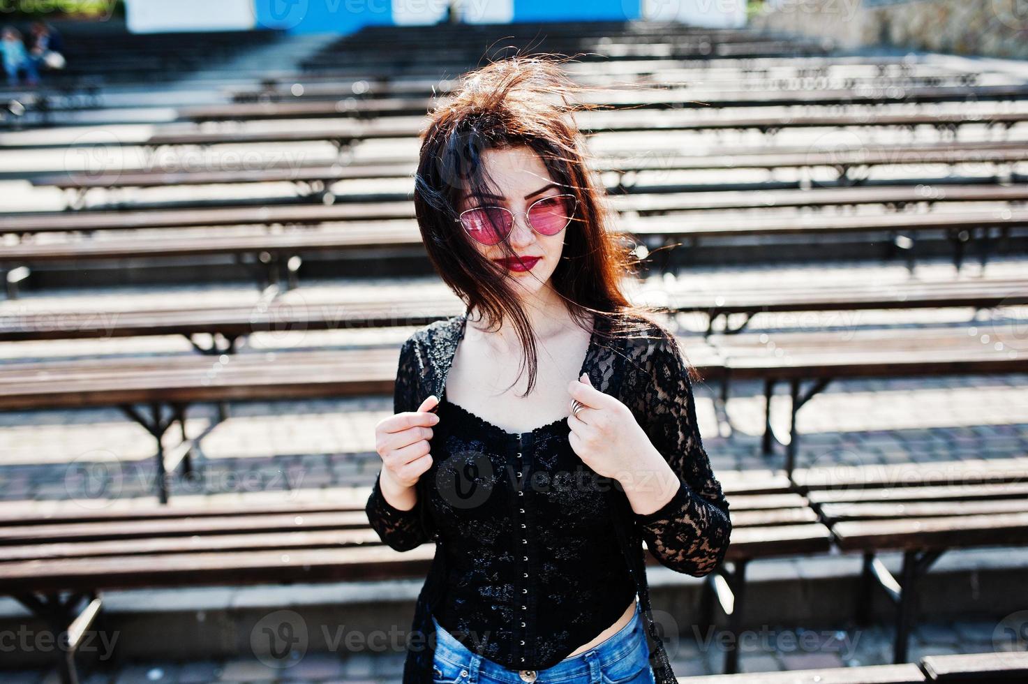 retrato de una chica morena con gafas rosas puestas en negro al aire libre en un día soleado contra una fila de bancos. foto