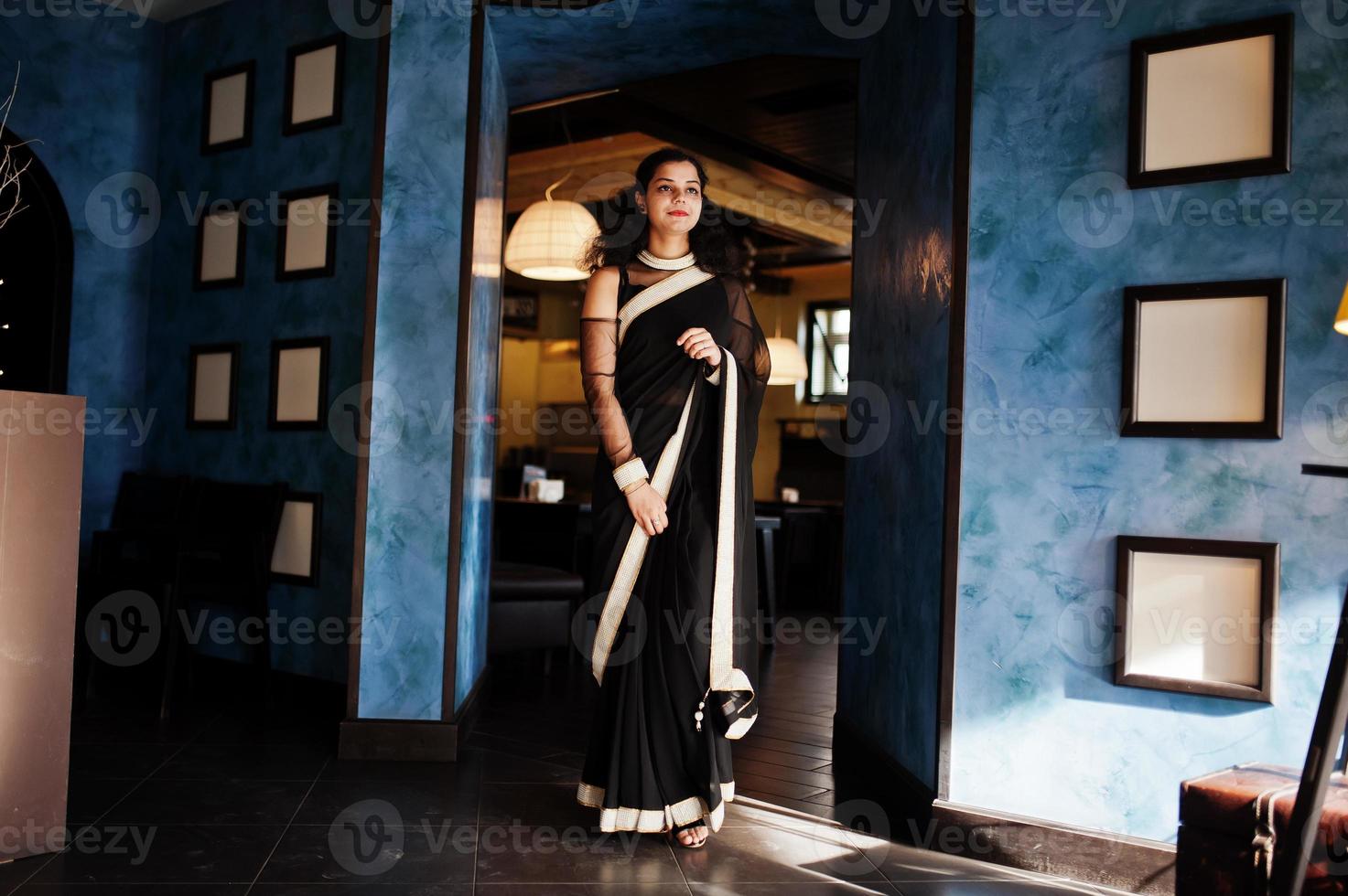 Young indian woman wear at elegant black saree posed on restaurant against wall with frames. photo