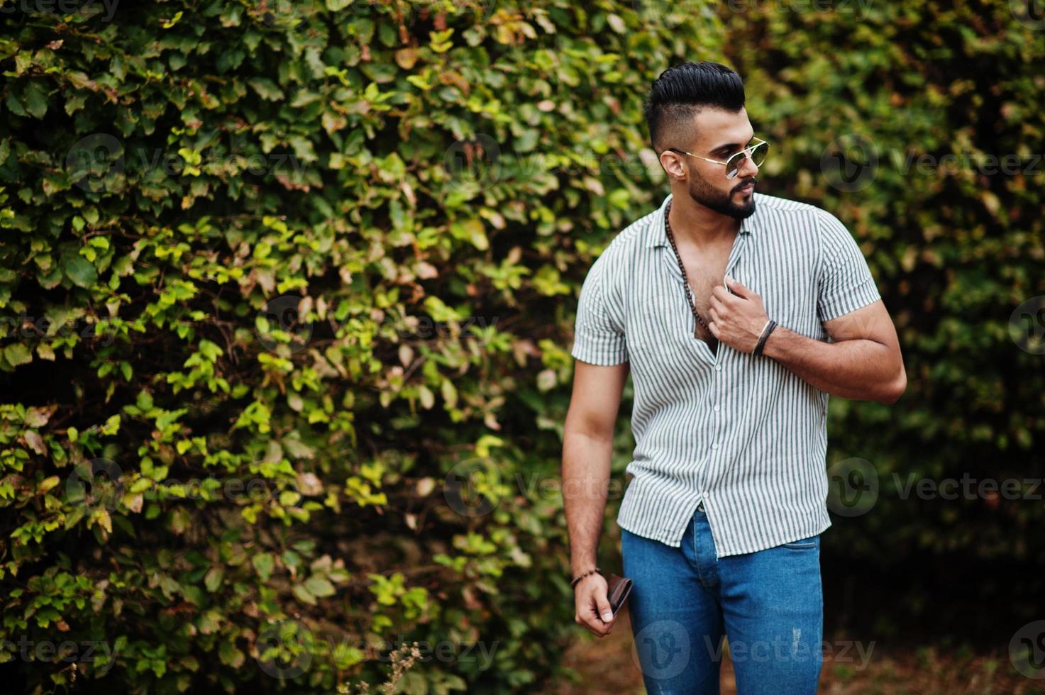 Fashionable tall arab beard man wear on shirt, jeans and sunglasses posed on park against greenery. photo