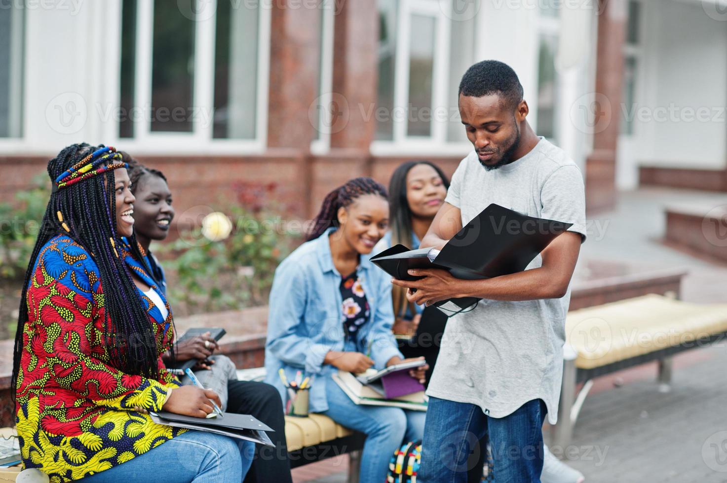 Group of five african college students spending time together on campus at university yard. Black afro friends studying at bench with school items, laptops notebooks. photo