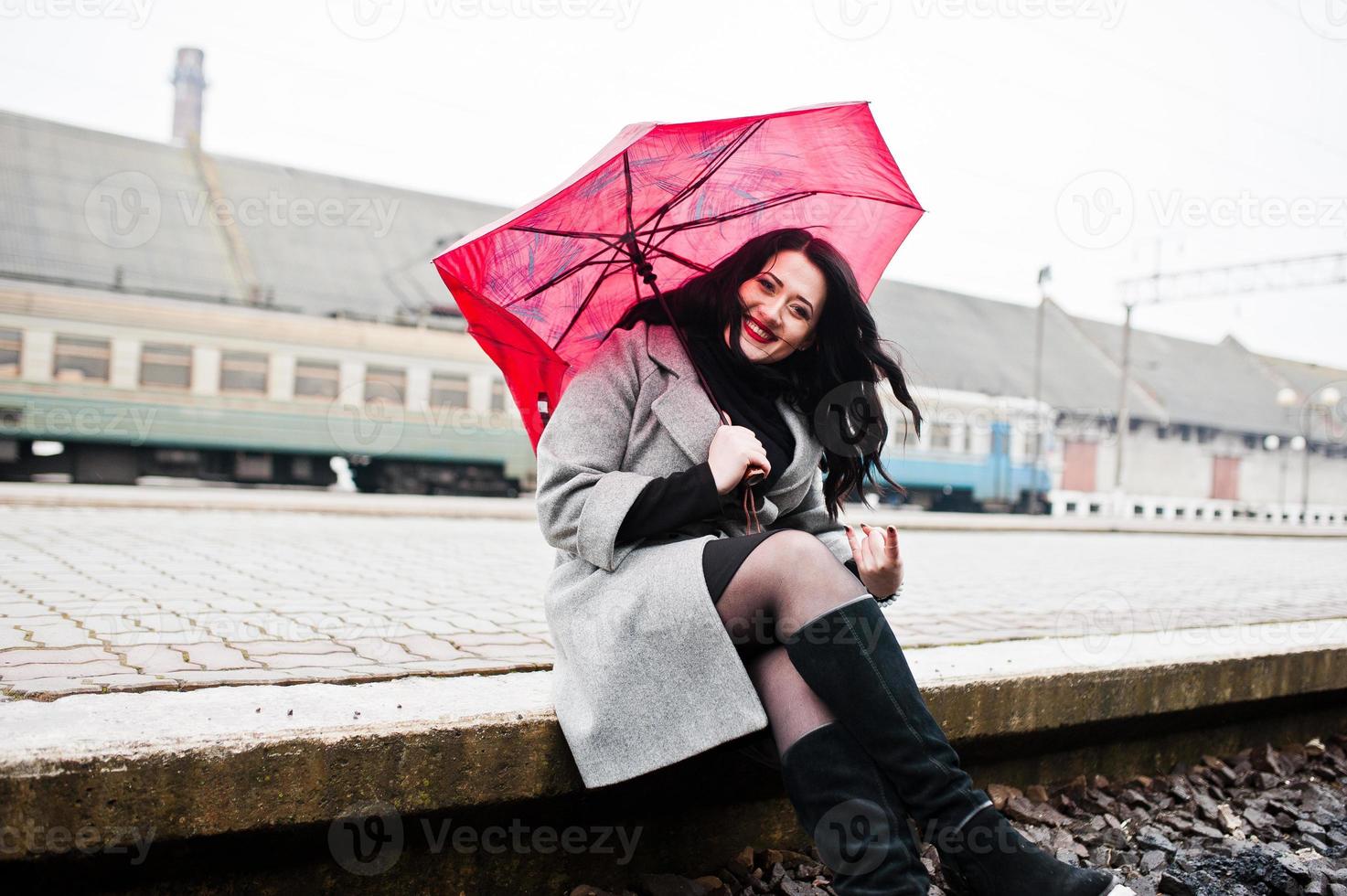 Brunette girl in gray coat with red umbrella in railway station. photo