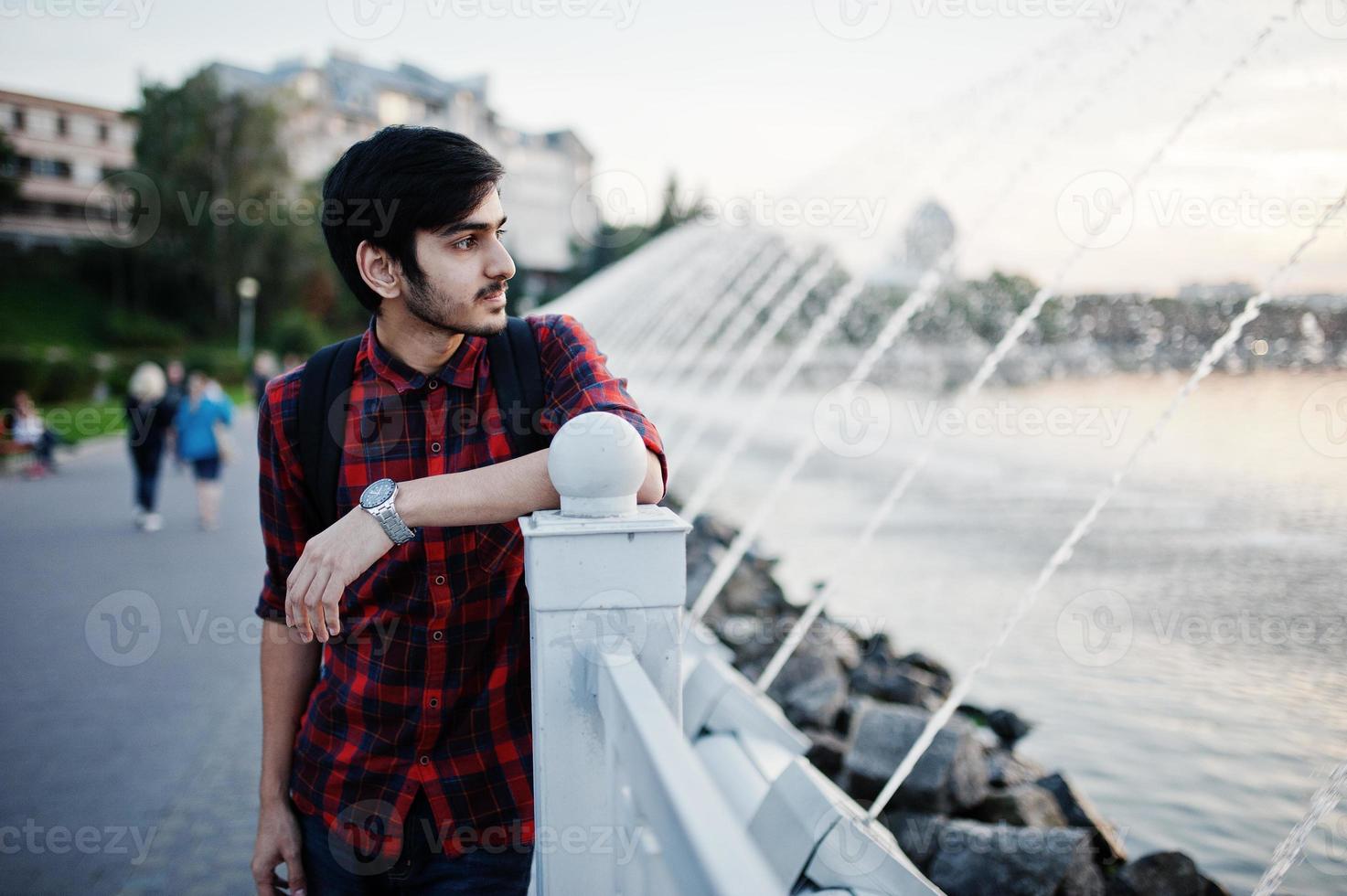 Young indian student man at checkered shirt and jeans with backpack posed on evening city against fountains. photo