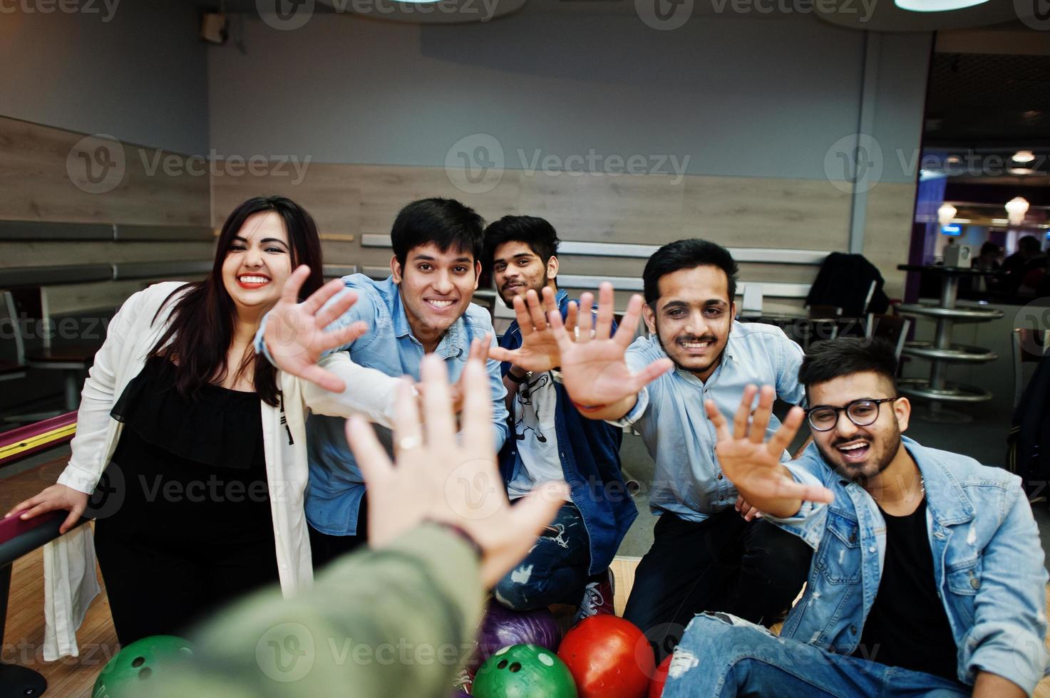 Group of five south asian peoples having rest and fun at bowling club. Holding bowling balls, sitting on alley and giving high five by hands. photo