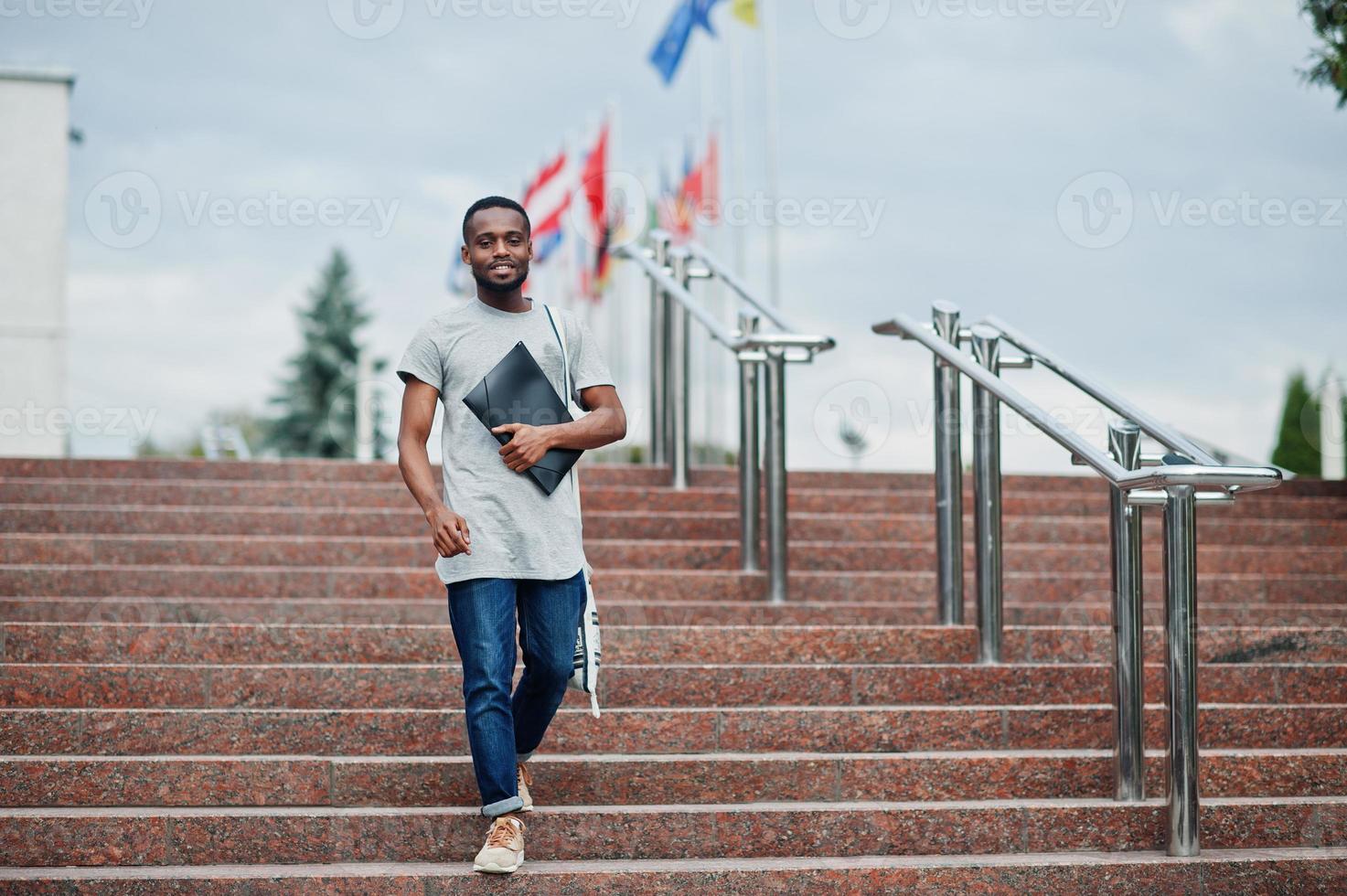 African student male posed with backpack and school items on yard of university, against flags of different countries. photo