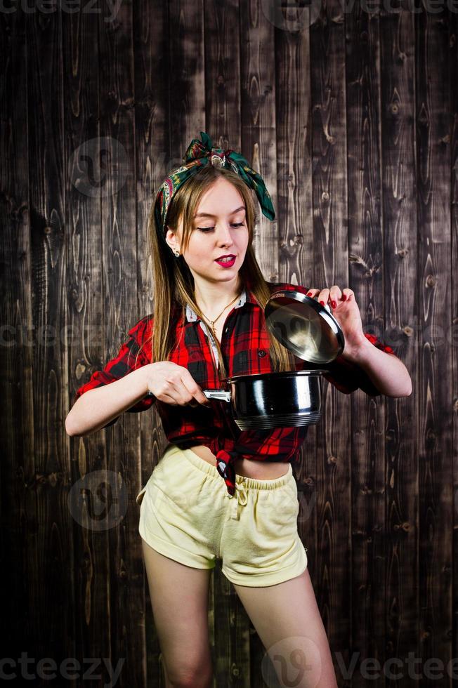 Young funny housewife in checkered shirt and yellow shorts pin up style with saucepan on wooden background. photo