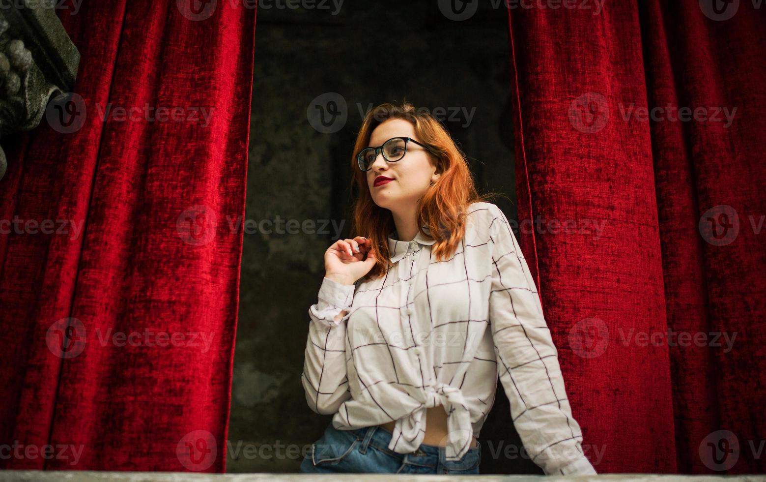 Attractive redhaired woman in eyeglasses, wear on white blouse posing at arch of open red curtains. photo