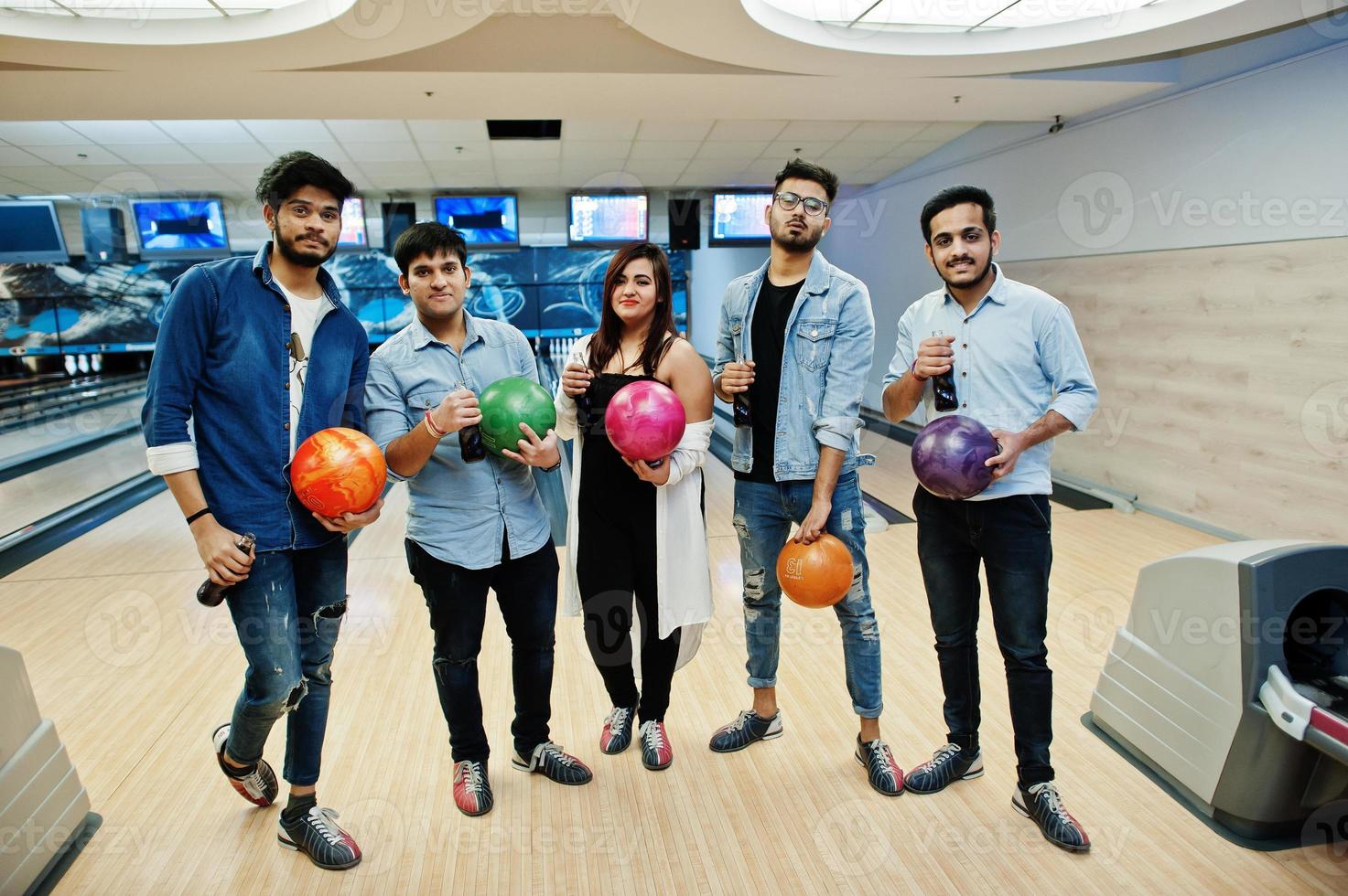 Group of five south asian peoples having rest and fun at bowling club. Holding cold soda drinks from glass bottles and bowling balls at hands. photo