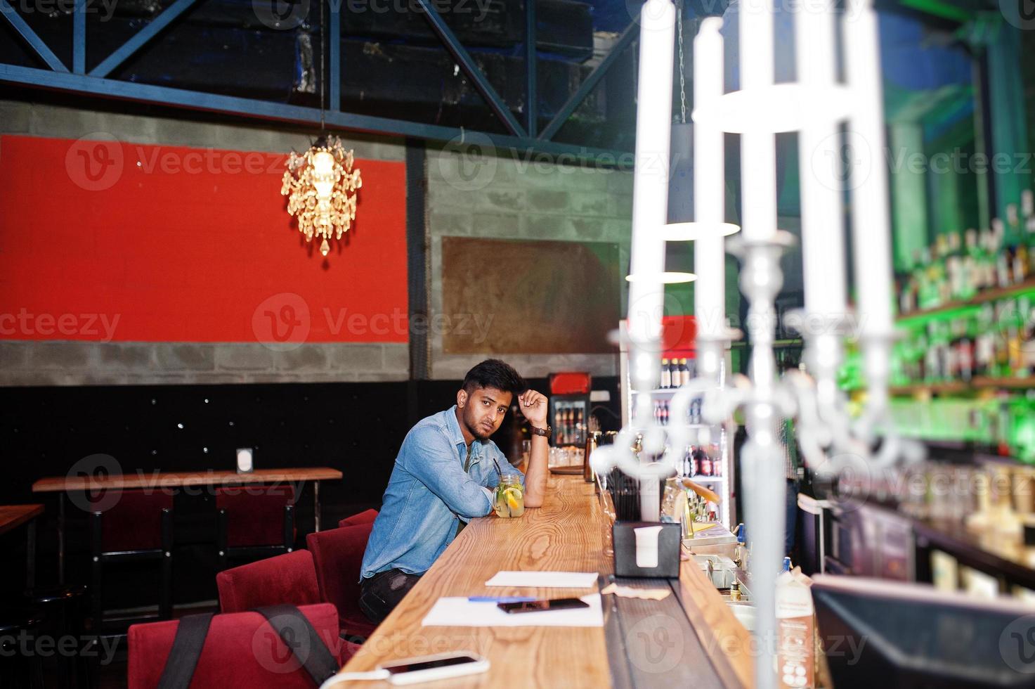 Portrait of handsome successful bearded south asian, young indian freelancer in blue jeans shirt sitting in night club against bar counter with cocktail and having a rest. photo