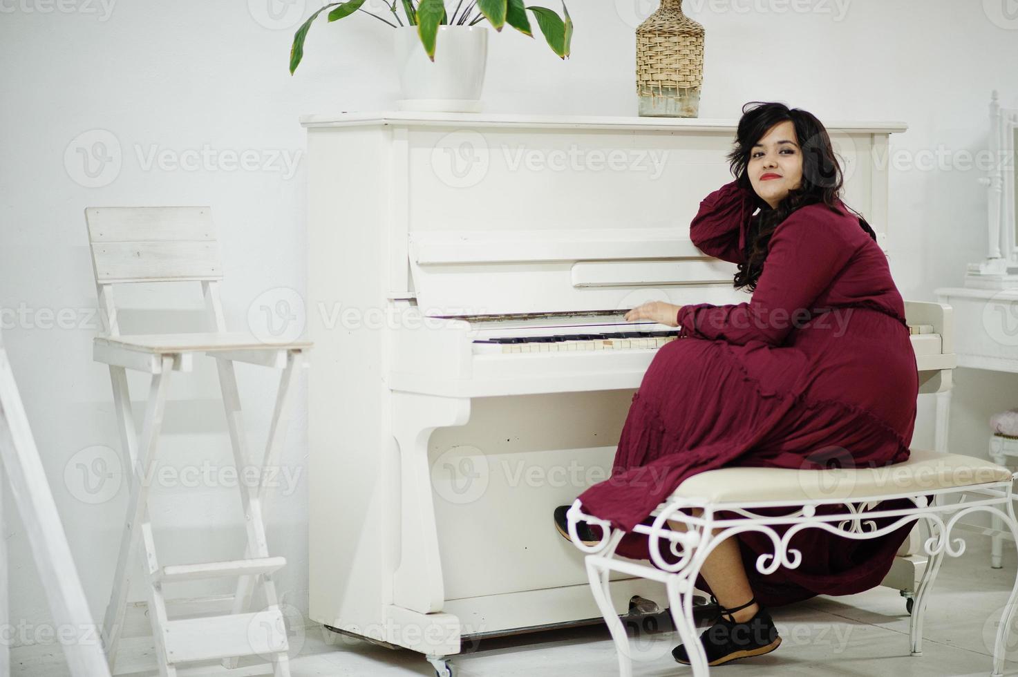 atractiva mujer del sur de asia con un vestido rojo intenso posó en la habitación y tocó el piano. foto