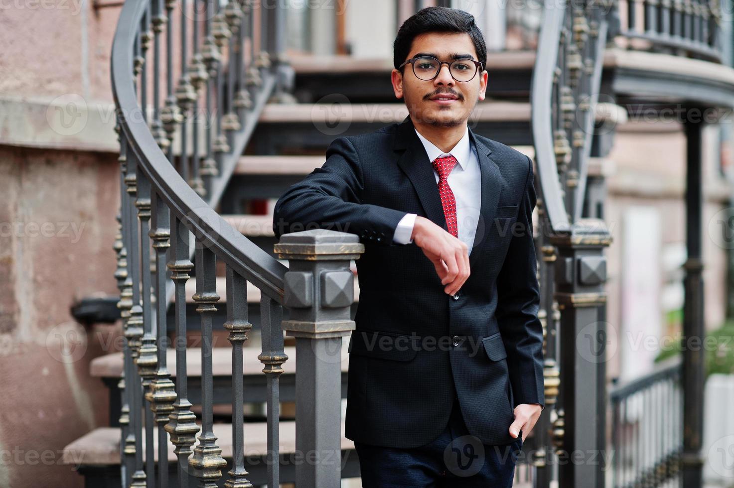 joven indio con anteojos, traje negro con corbata roja posado al aire libre contra escaleras de hierro. foto