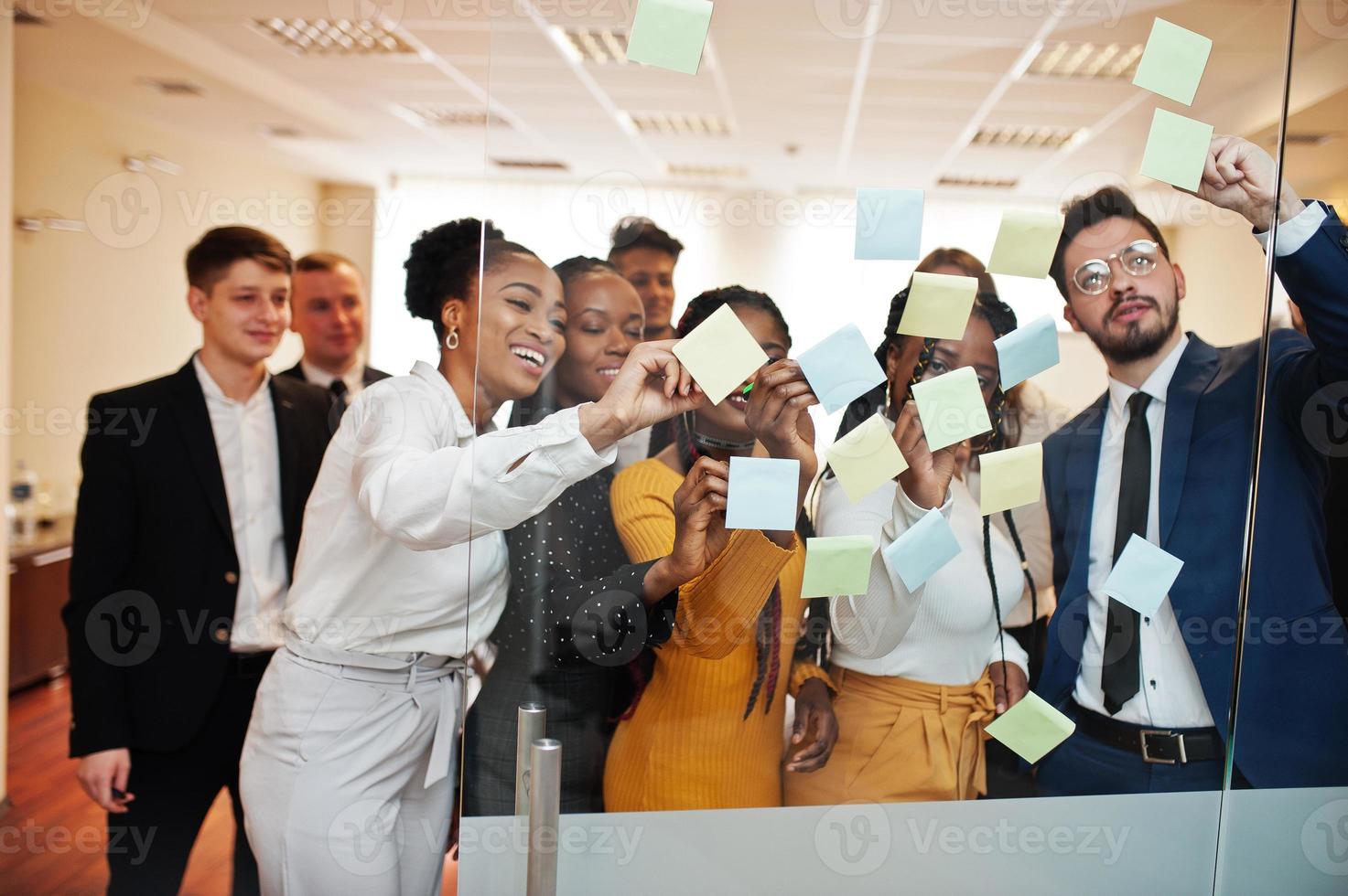 Team of multicultural young people pointing on glass with colorful paper notes. Diverse group of male and female employees in formal wear using stickers. photo