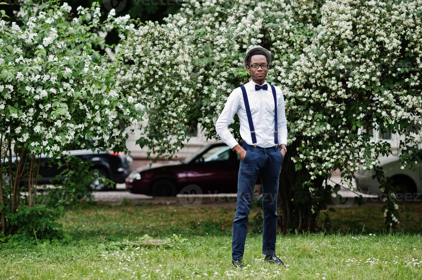 Stylish african american business man at pants with suspender and shirt with bow tie, hat and glasses posed outdoor. photo