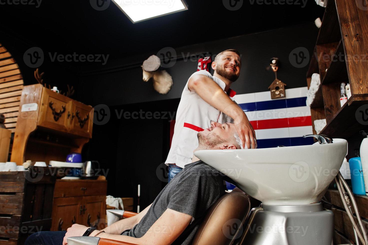 Young bearded man washing head by hairdresser while sitting in chair at barbershop. Barber soul. photo