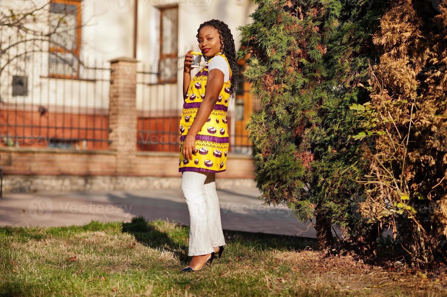 Stylish african american women in yellow jacket posed on street with hot drink in disposable paper cup. photo