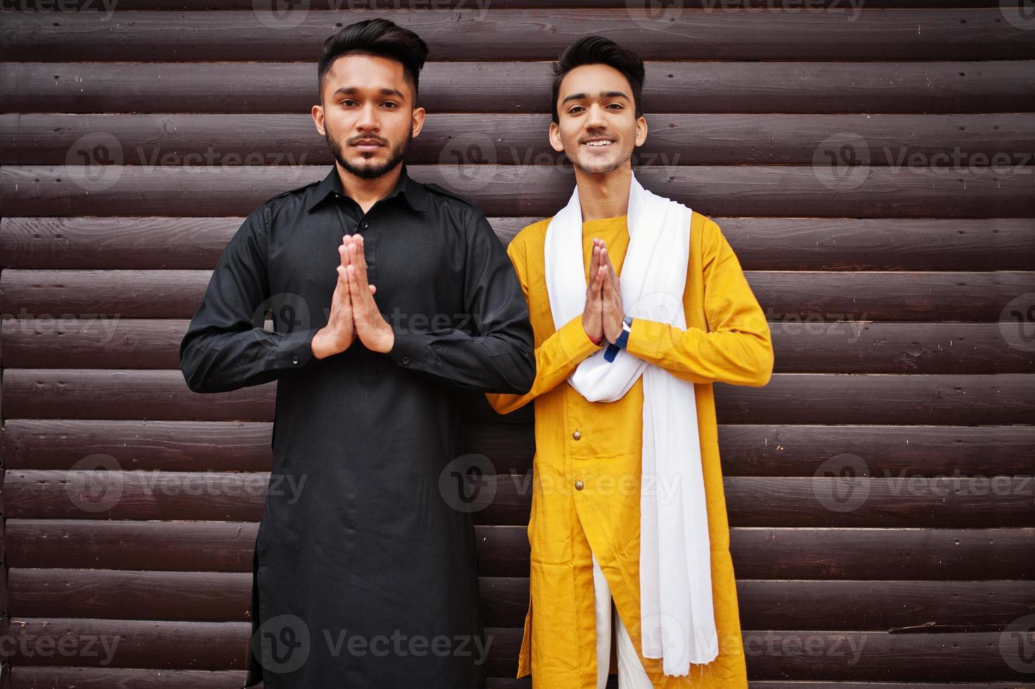 Two indian stylish mans friends in traditional clothes posed outdoor against wooden background and shows namaste hands sign. photo