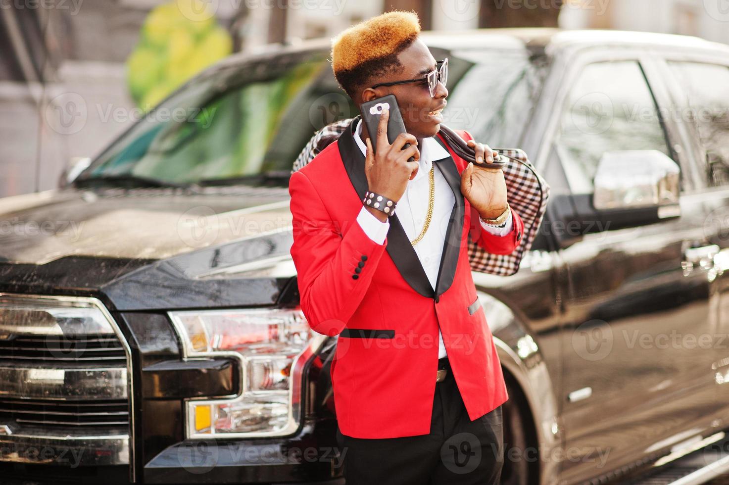 Fashion african american man model at red suit, with highlights hair posed against big suv black car and speaking on mobile phone. photo