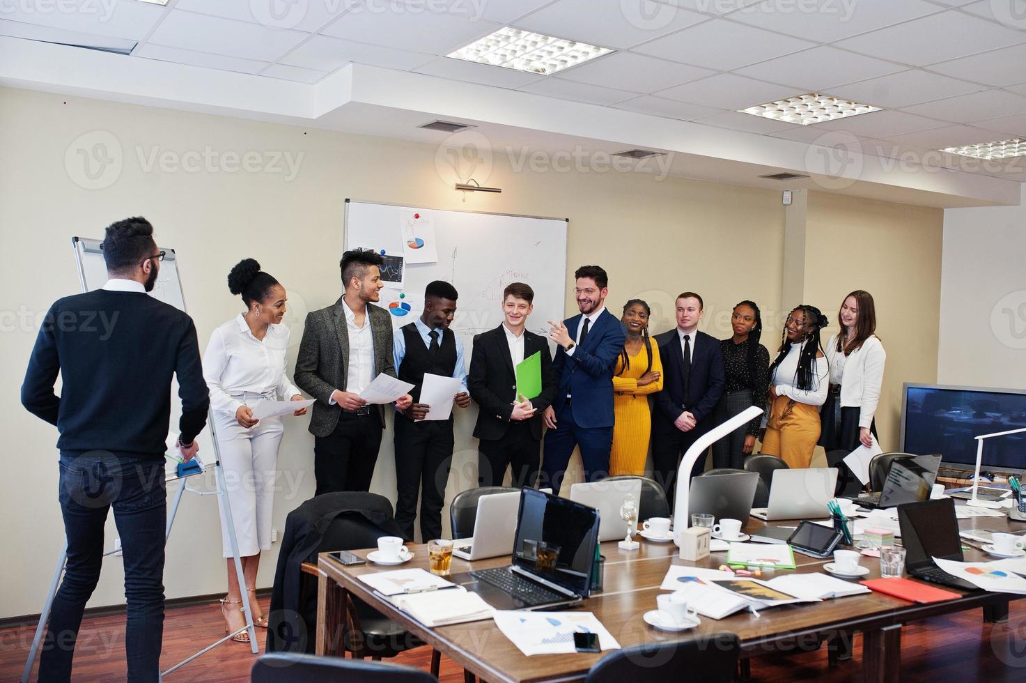 Mixed race business coach presenting report standing near whiteboard pointing on sales statistic shown on diagram and chart teach diverse company members gathered together in conference room. photo