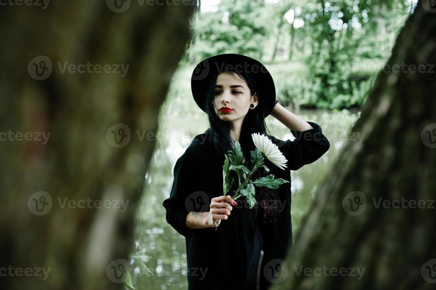 chica sensual todo en negro, labios rojos y sombrero. mujer gótica dramática sostiene una flor de crisantemo blanco a mano contra el lago silencioso. foto