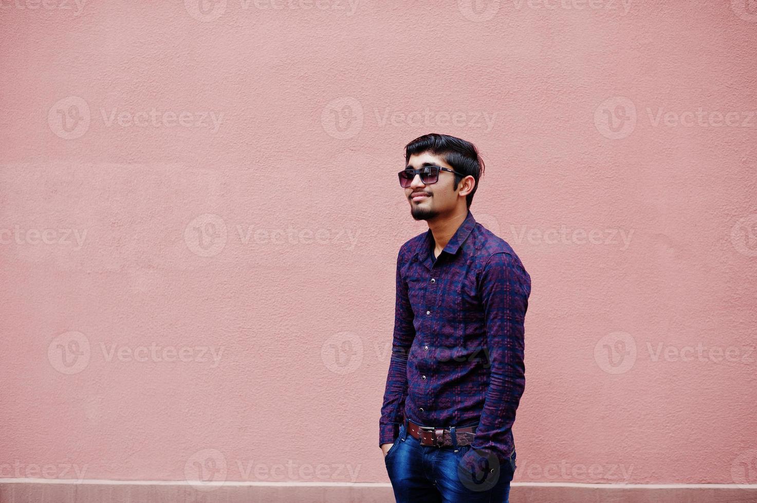 Young indian man on shirt and sunglasses posed against pink wall. photo