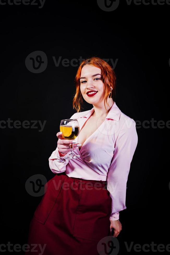 Gorgeous red haired girl in pink blouse and red skirt with glass of wine at hand isolated on black. photo