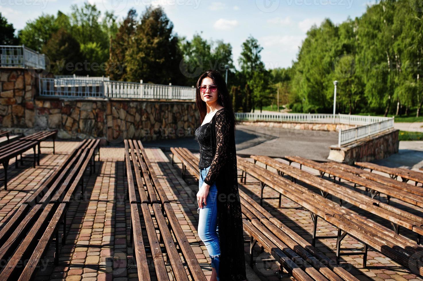 Portrait of brunette girl in pink glasses wear on black posed outdoor on sunny day against row of benches. photo