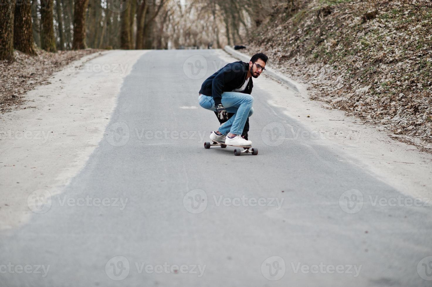 Street style arab man in eyeglasses with longboard longboarding down the road. photo