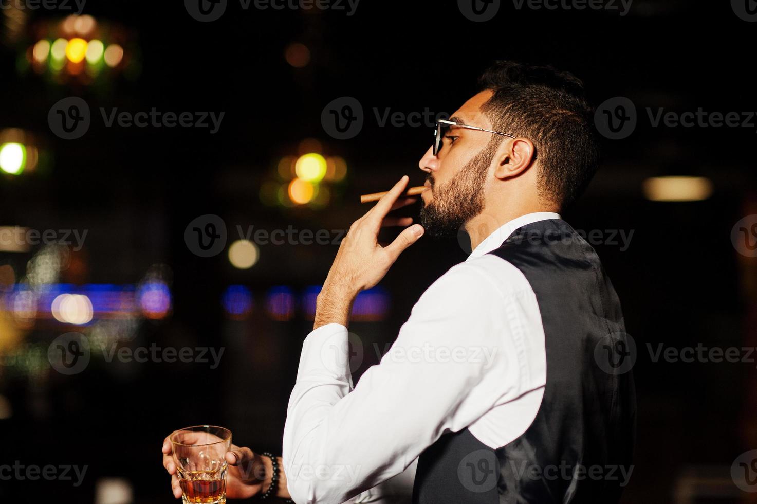 apuesto hombre árabe bien vestido con vaso de whisky y cigarro posado en el pub. foto