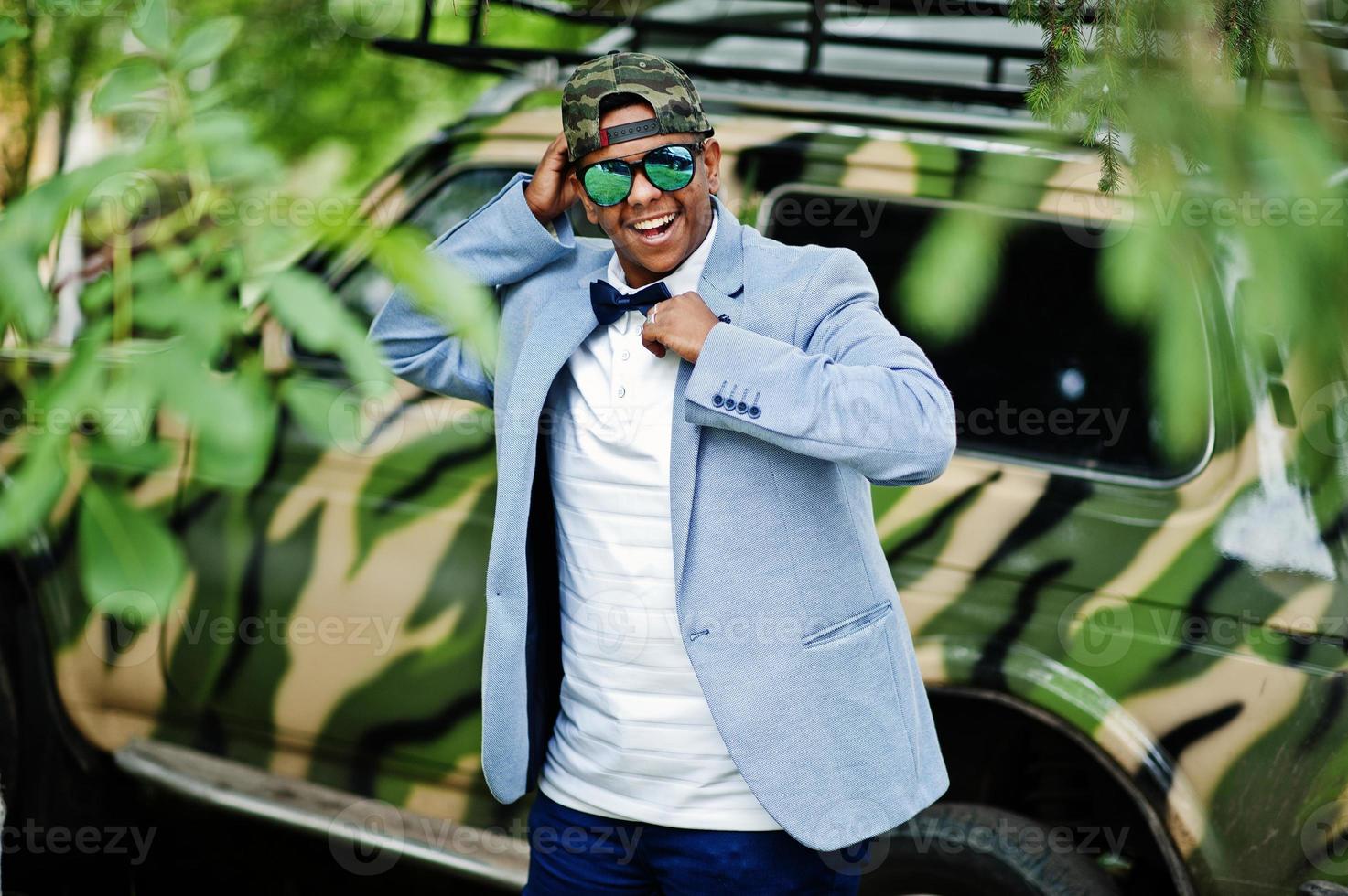 elegante hombre árabe con gafas de sol, gorra y corbata de moño posado contra un coche militar. chico modelo árabe. foto