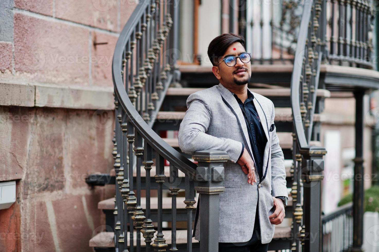hombre indio elegante con bindi en la frente y anteojos, traje puesto al aire libre contra escaleras de hierro. foto