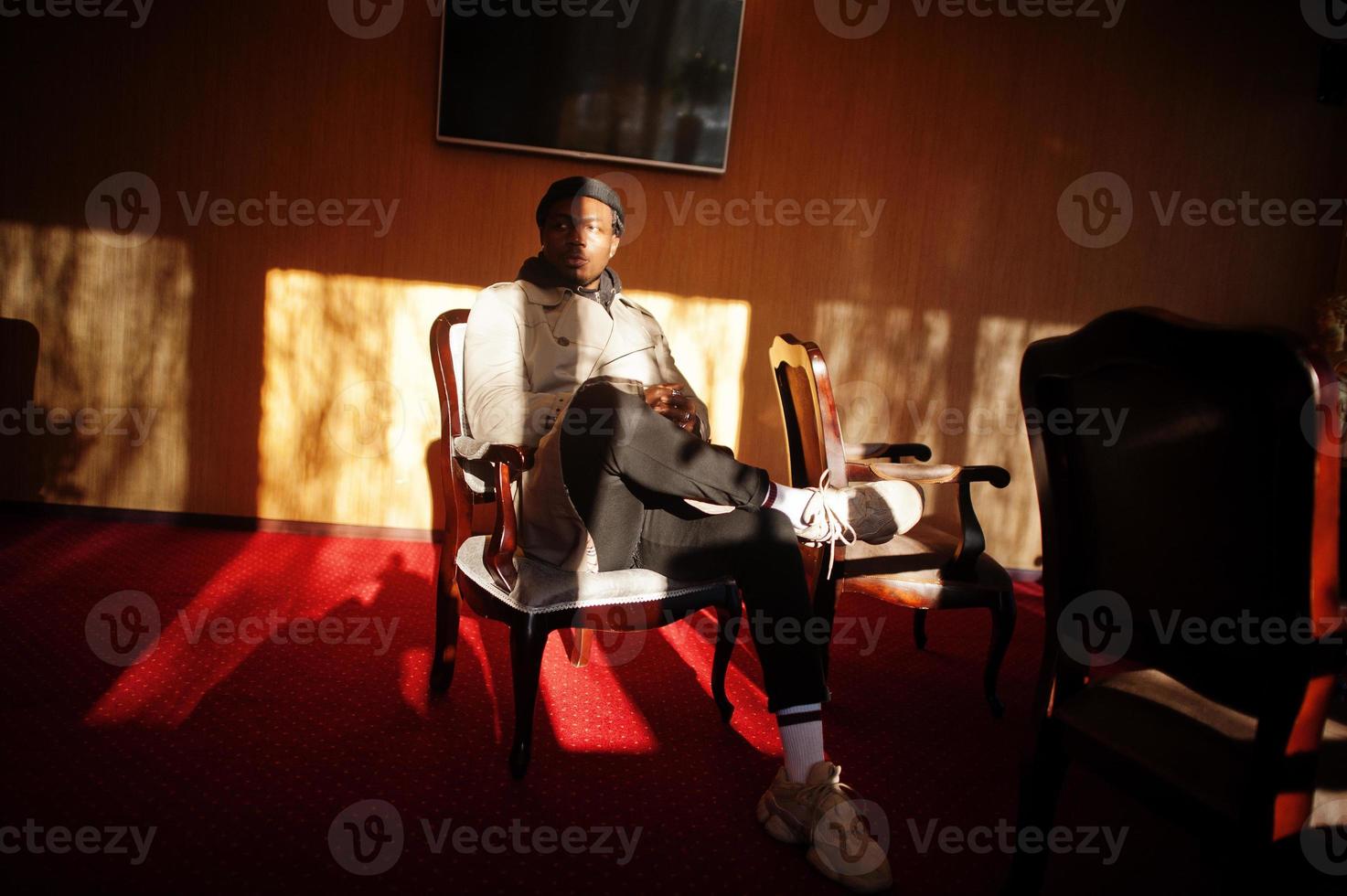 apuesto hombre afroamericano posando dentro de la habitación con sombras de luz solar en sombrero negro y abrigo beige. foto
