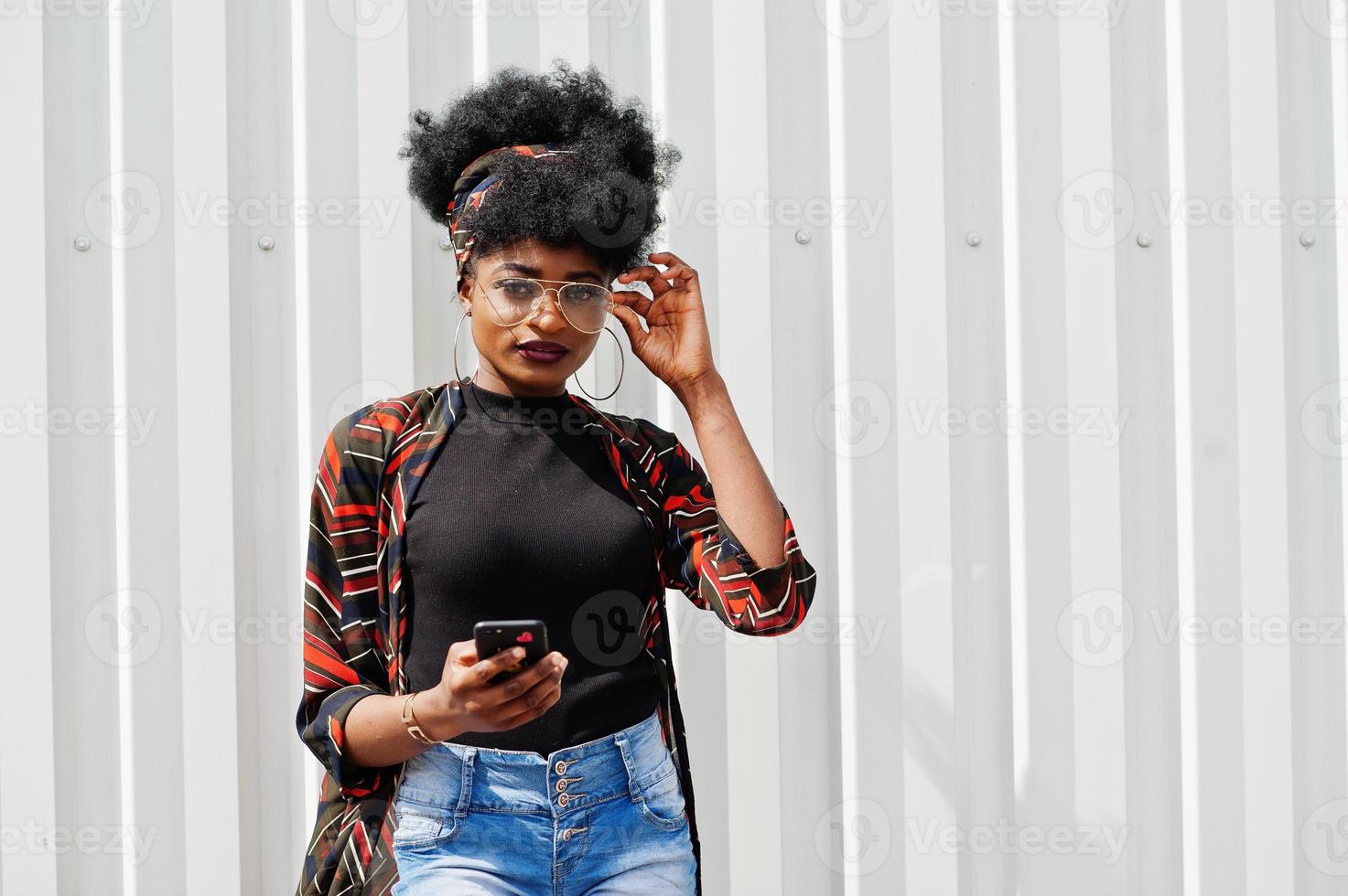 African woman with afro hair, in jeans shorts and eyeglasses posed against white steel wall with mobile phone in hand. photo