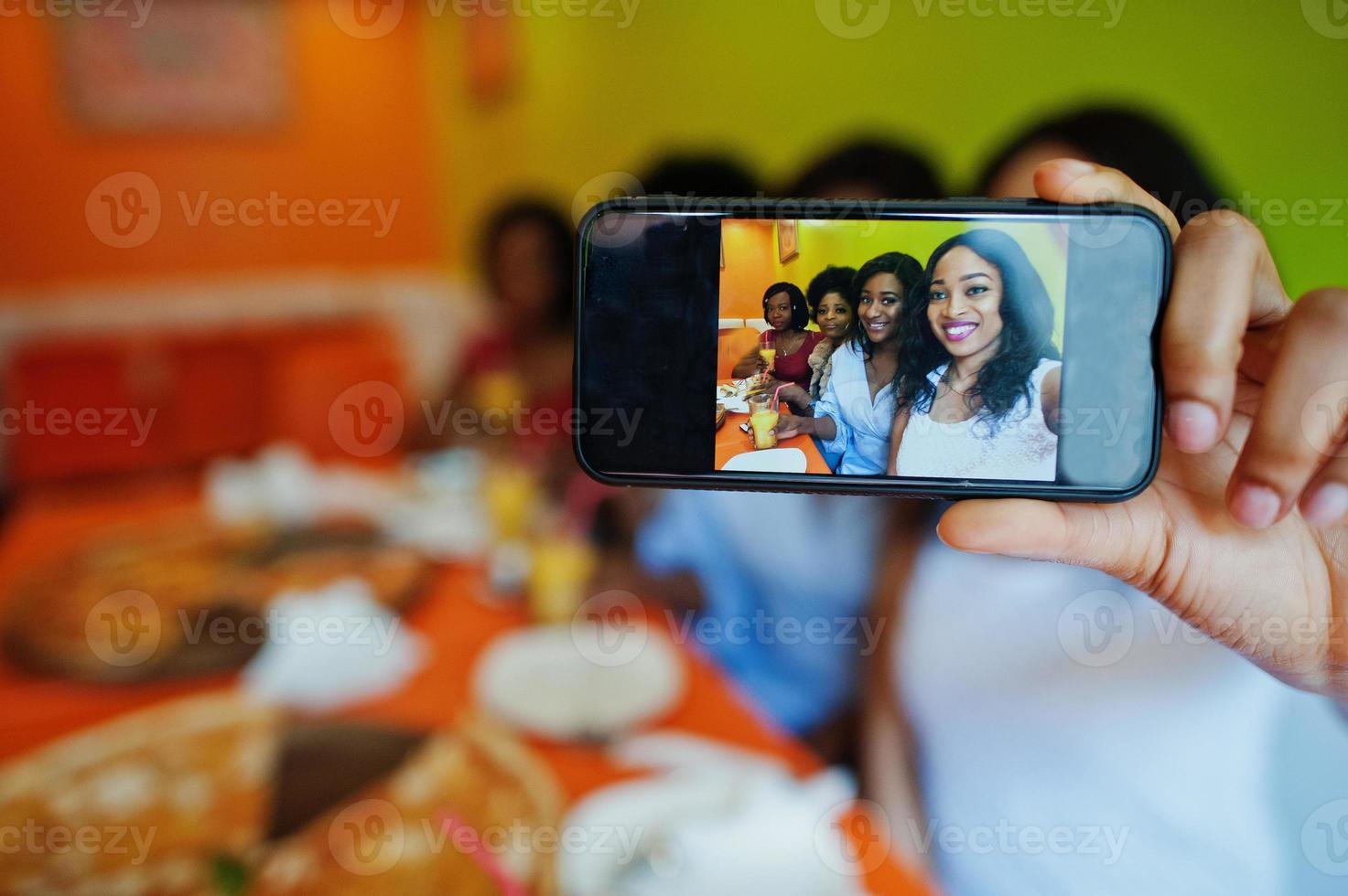 cierre la pantalla del teléfono de fotos de cuatro jóvenes africanas en un restaurante de colores brillantes comiendo pizza, divirtiéndose juntas y haciendo selfie.