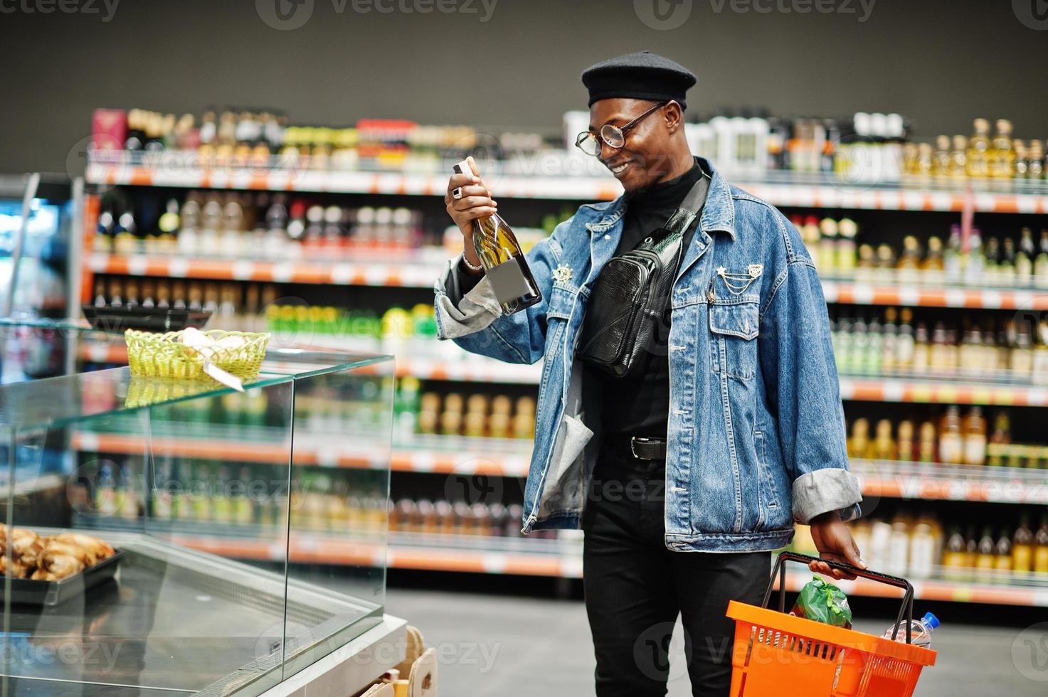 elegante hombre afroamericano casual con chaqueta de jeans y boina negra sosteniendo una canasta y mirando una botella de vino, comprando en el supermercado. foto