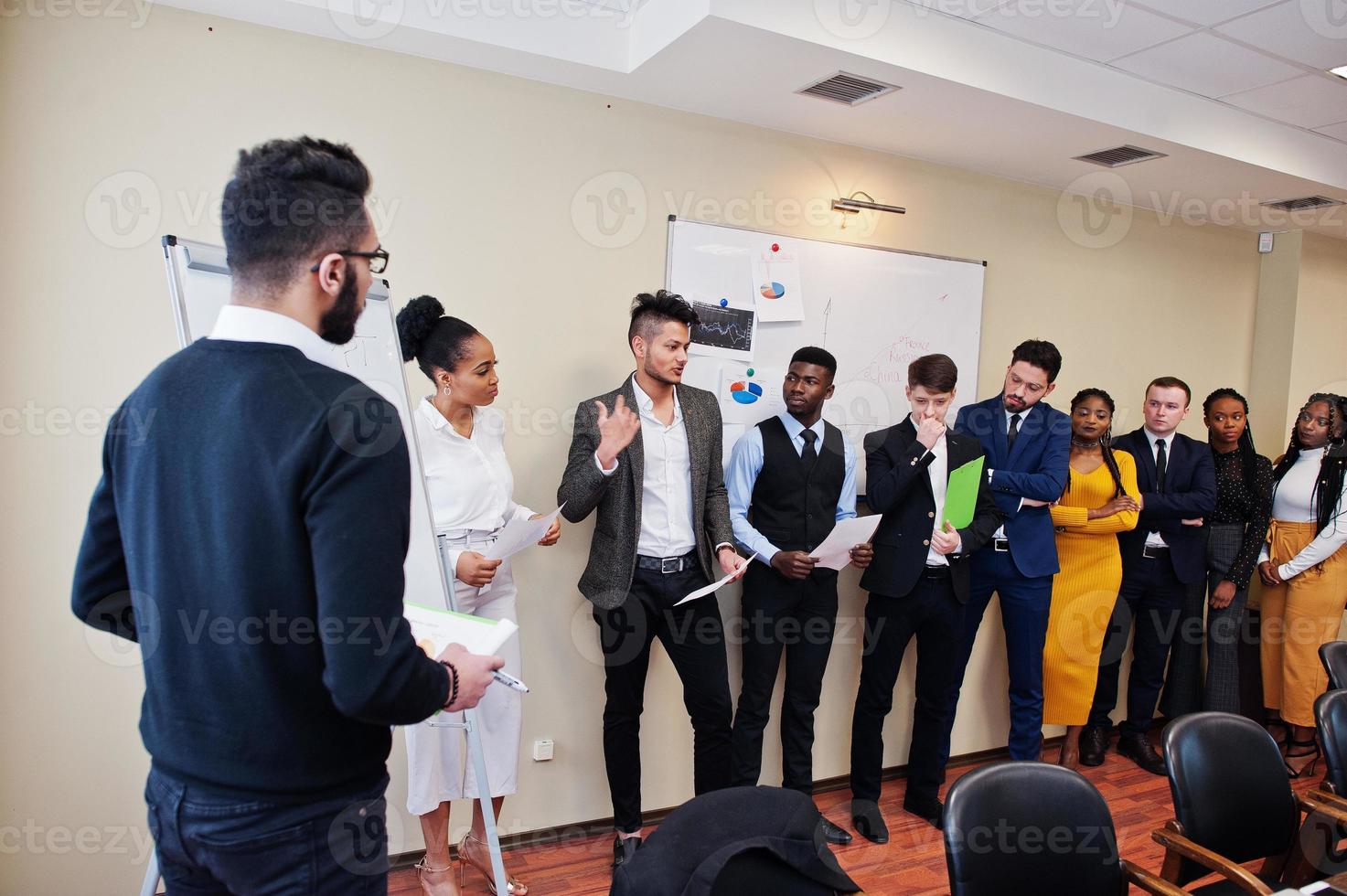 Mixed race business coach presenting report standing near whiteboard pointing on sales statistic shown on diagram and chart teach diverse company members gathered together in conference room. photo