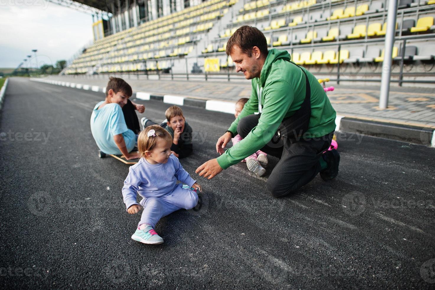 cuatro niños con padre en asfalto juegan y se divierten. la familia deportiva pasa tiempo libre al aire libre con scooters y patines. foto