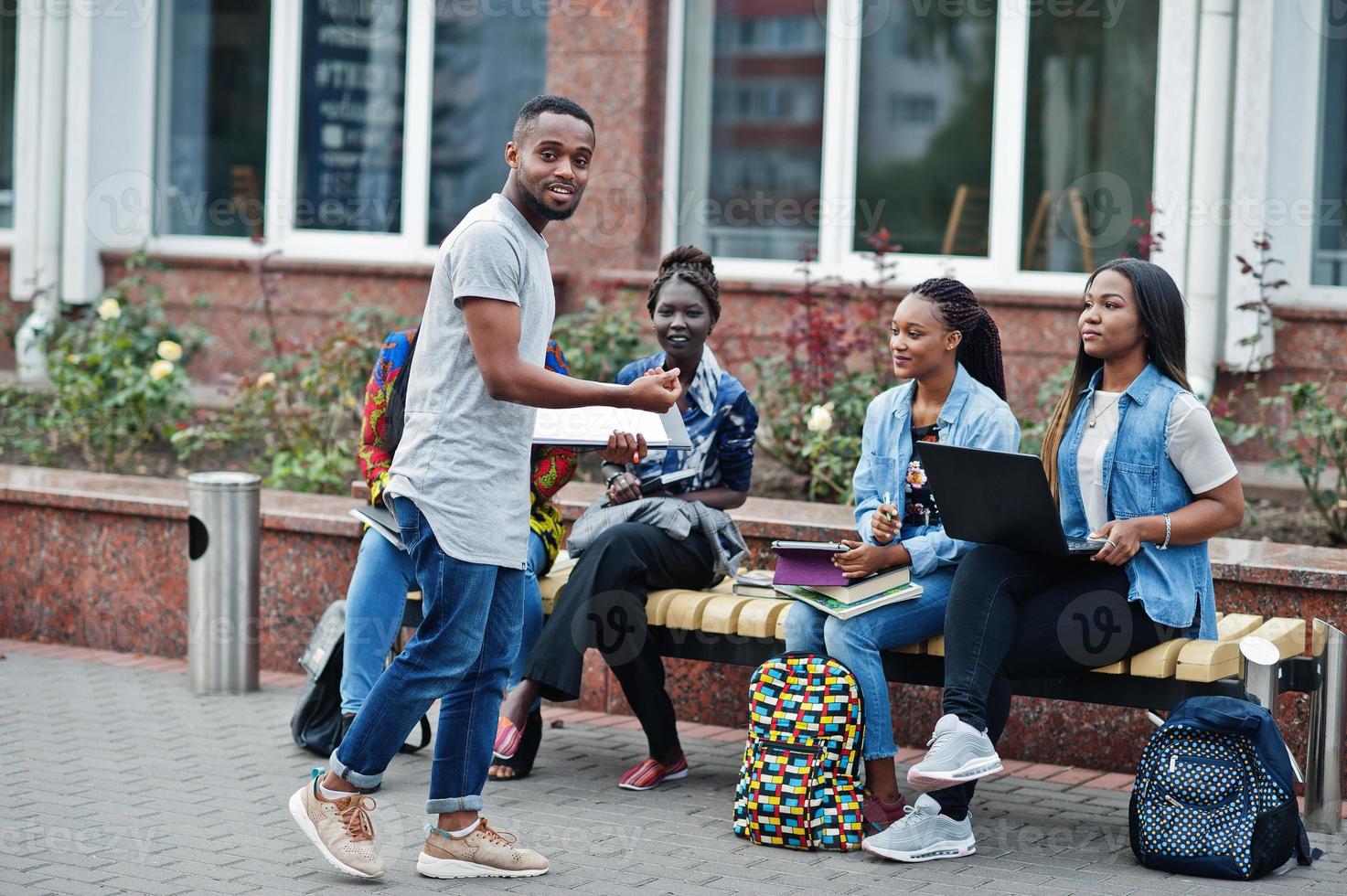 Group of five african college students spending time together on campus at university yard. Black afro friends studying at bench with school items, laptops notebooks. photo