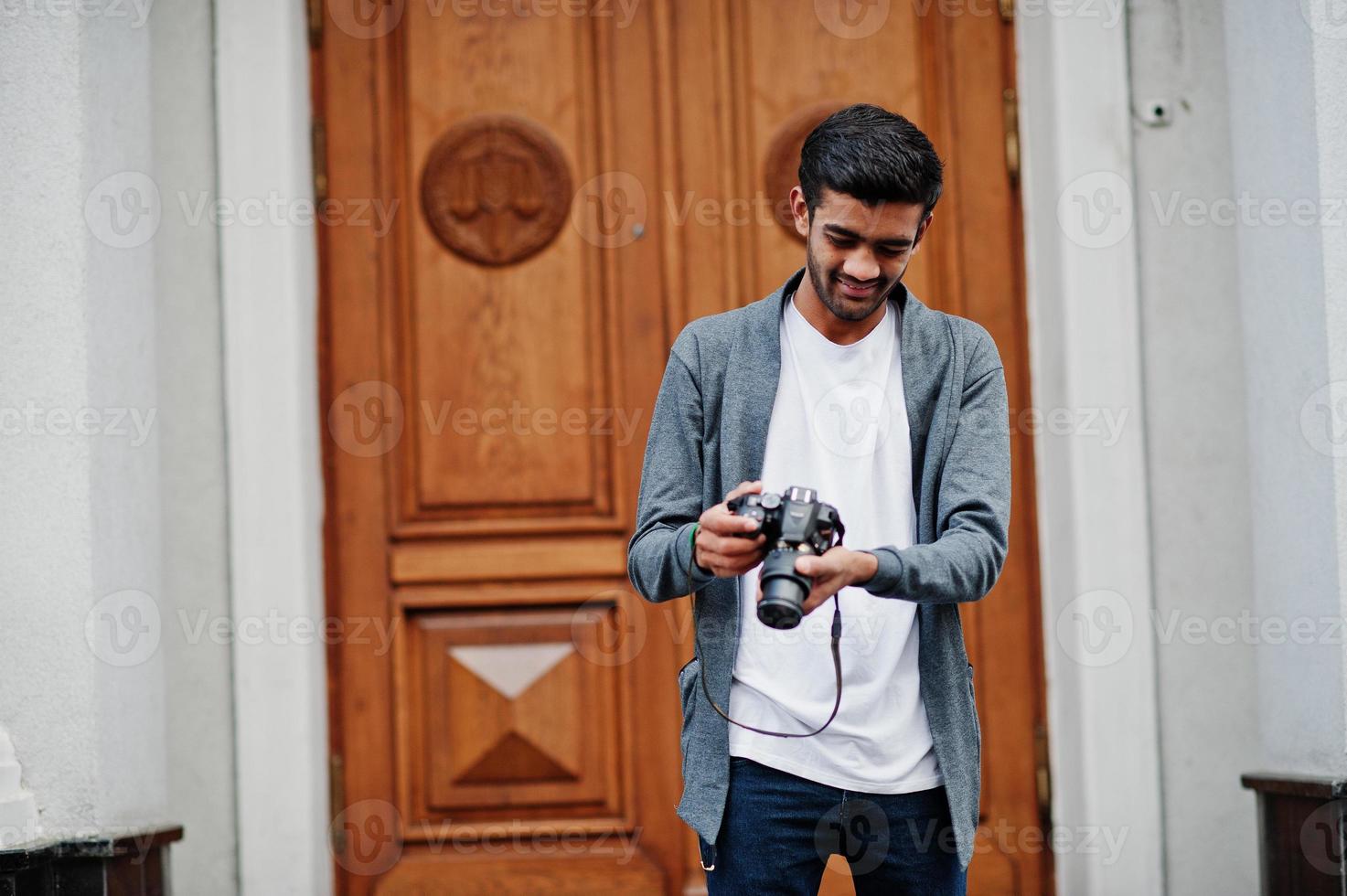 Un joven fotógrafo indio con estilo viste una pose informal al aire libre con una cámara fotográfica dslr en las manos. foto