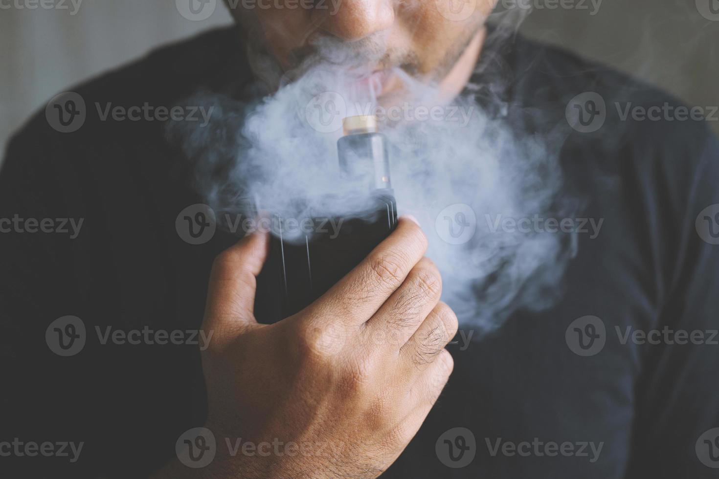 photo portrait young man with beard hold and smoking his electronic cigarette blowing a stream of smoke spread. dark black, grey background