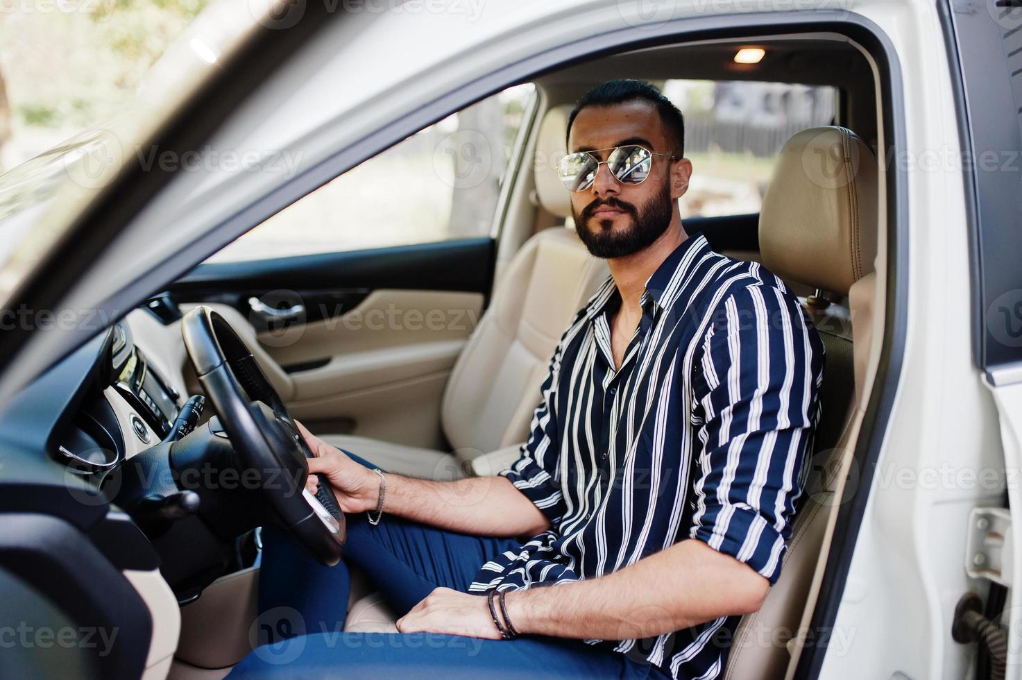 Successful arab man wear in striped shirt and sunglasses pose behind the wheel of  his white suv car. Stylish arabian men in transport. photo