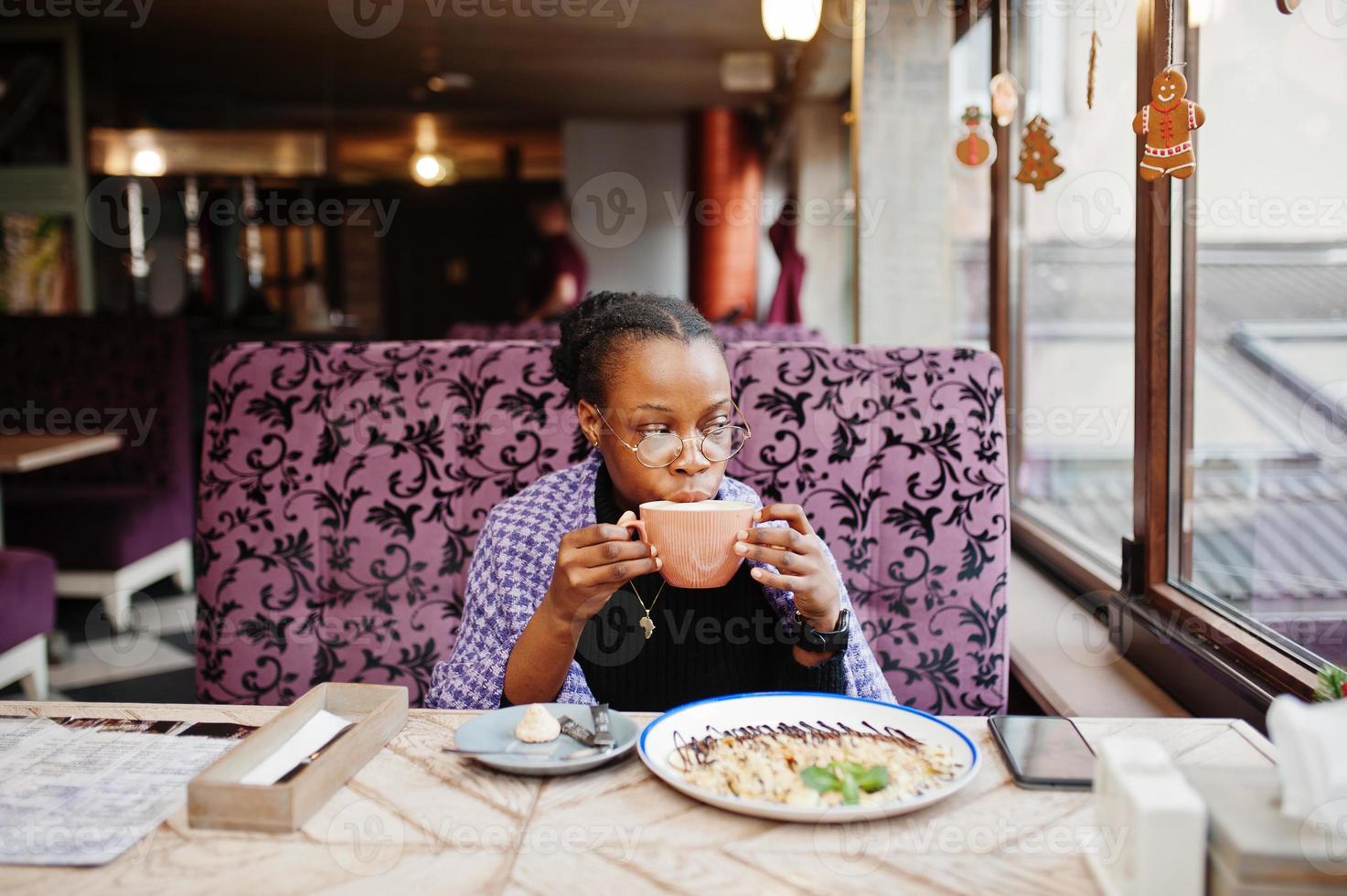 African woman in checkered violet cape and eyeglasses posed at cafe, sitting by table with dessert and cup of coffee. photo