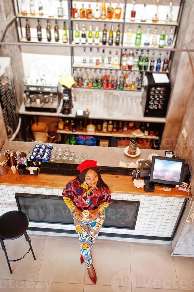 Enthusiastic african american woman in trendy coloured outfit with red beret chilling in cozy cafe, standing near bar counter. photo