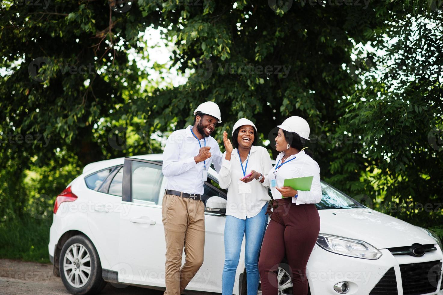 African american technician in white helmets near car. Group of three black engineers meeting. photo