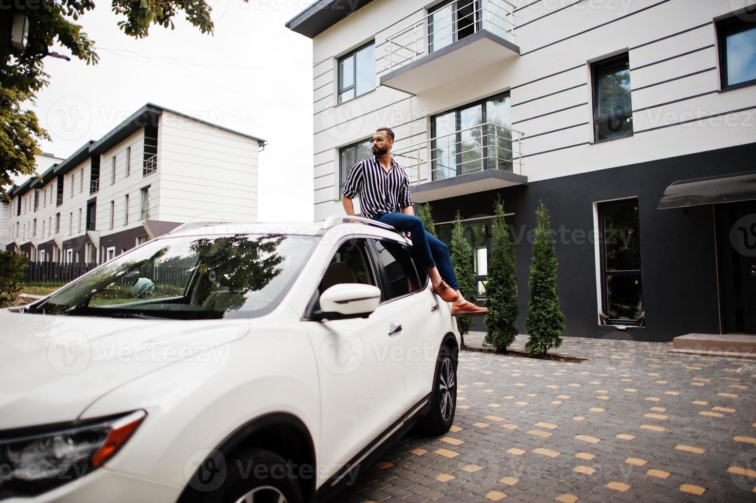 Successful arab man wear in striped shirt and sunglasses sitting on the roof of his white suv car. photo