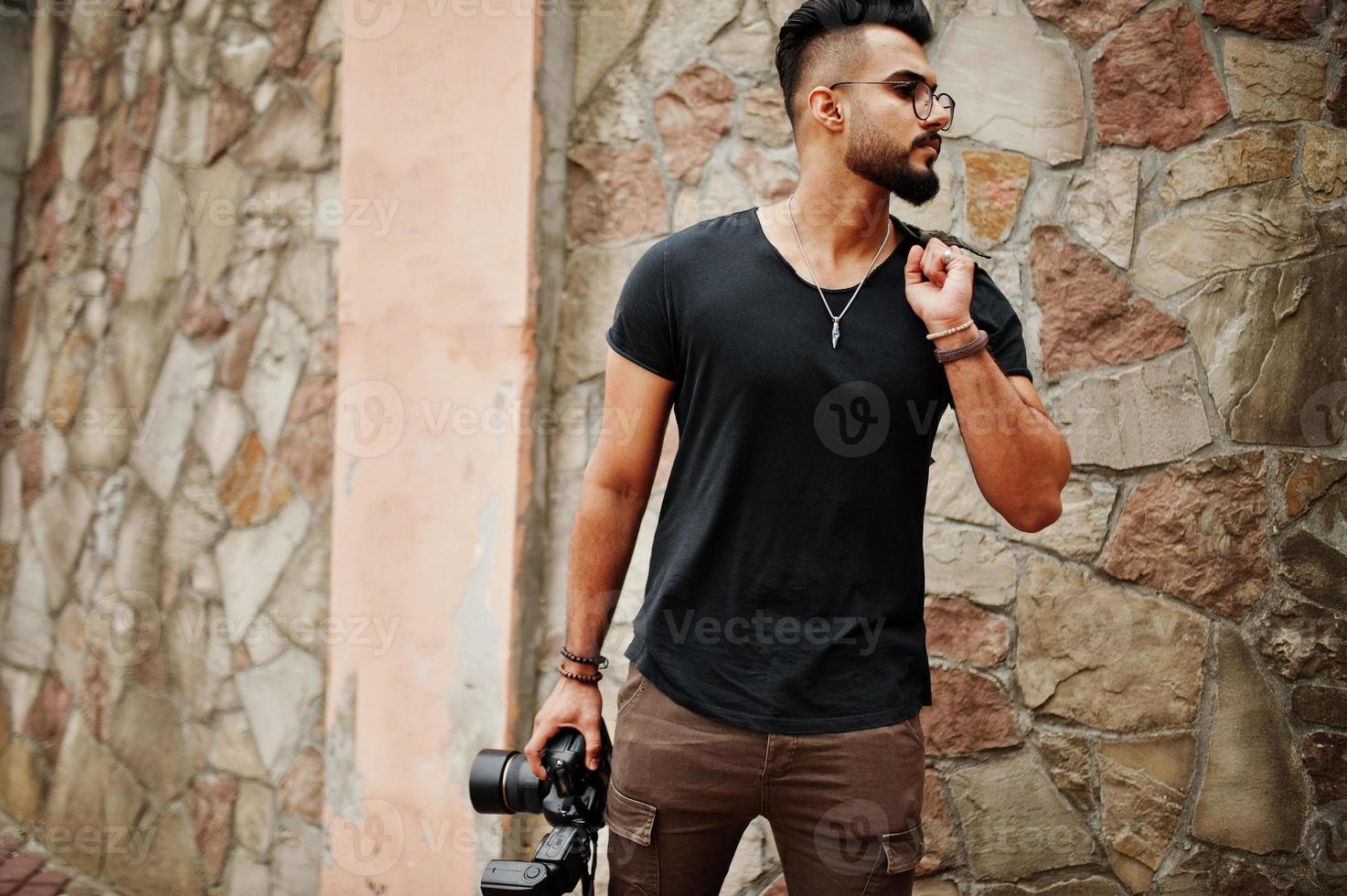 Impresionante y hermoso fotógrafo de hombre macho con barba árabe alta con gafas y camiseta negra con cámara profesional en las manos. foto