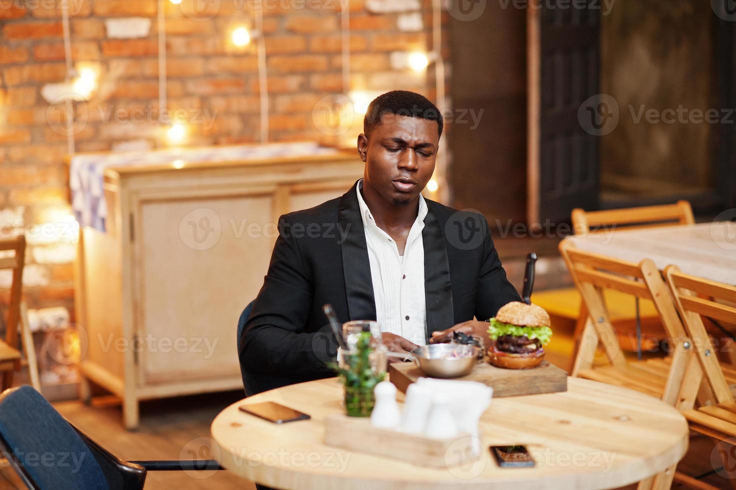 Respectable young african american man in black suit sitting in restaurant with tasty double burger and soda drink. photo