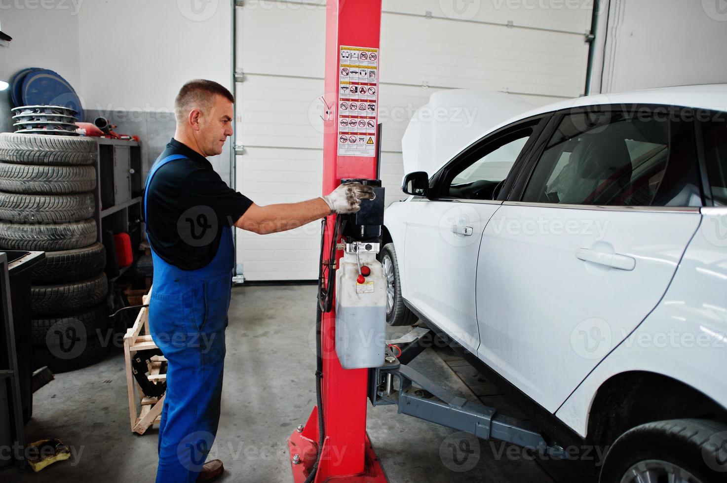 Car repair and maintenance theme. Mechanic in uniform working in auto service, press button to lifting auto. photo