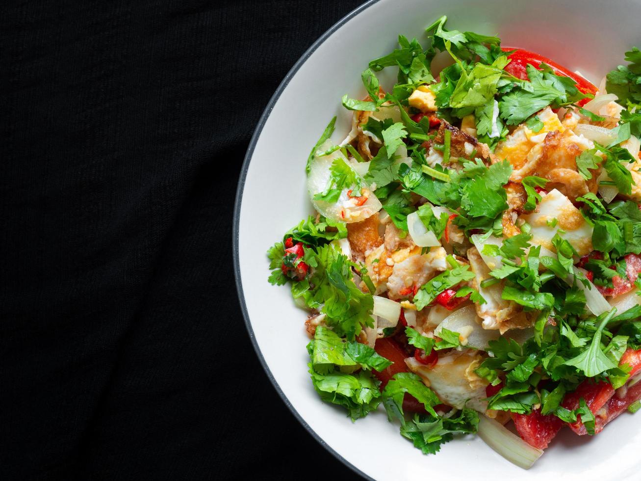 ensalada de huevo frito picante en un tazón blanco con fondo de tela negra menú de comida callejera comido popularmente por los tailandeses los ingredientes principales son huevos, cilantro, tomates, cebollas, agrio, salado. Puedes encontrarlo foto