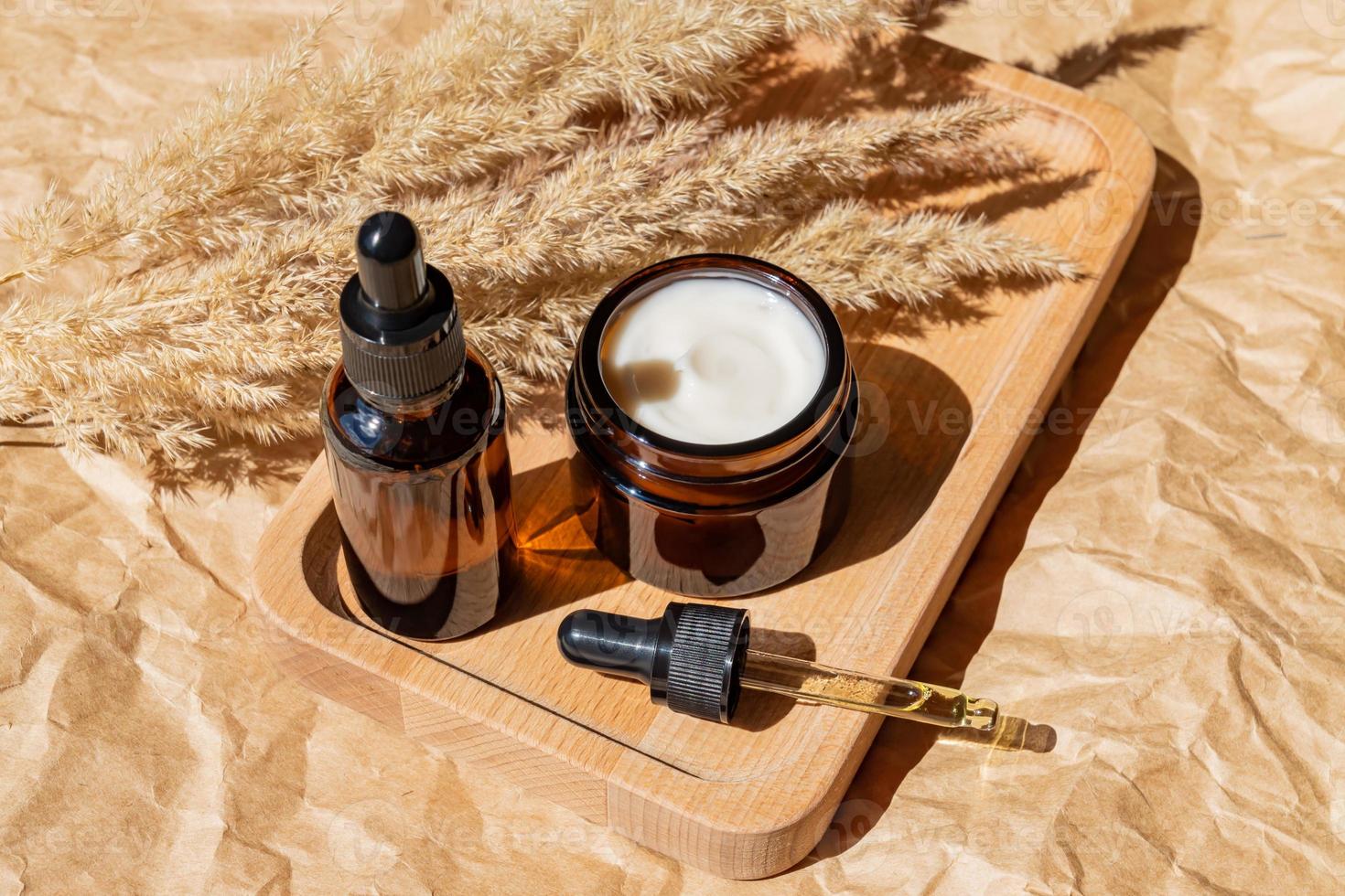 Blank amber glass essential oil bottle with pipette and face collagen cream on beige background decorated dry wild reeds. Skin care concept with natural cosmetics photo